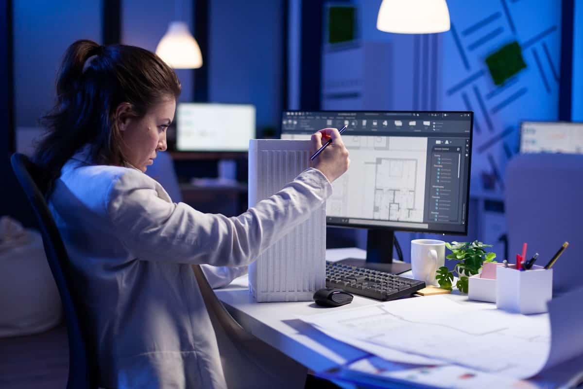 Woman architect analysing and matching blueprints for new building project sitting at desk. Graphic designer using arhitecture prototype plans working overtime in corporate office late at night